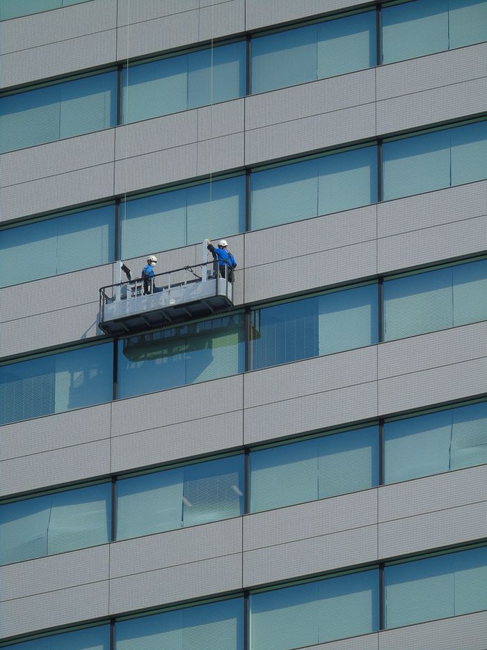 who-we-are Deux travailleurs nettoient les vitres de la façade d’un immeuble moderne en verre, depuis une plateforme suspendue.